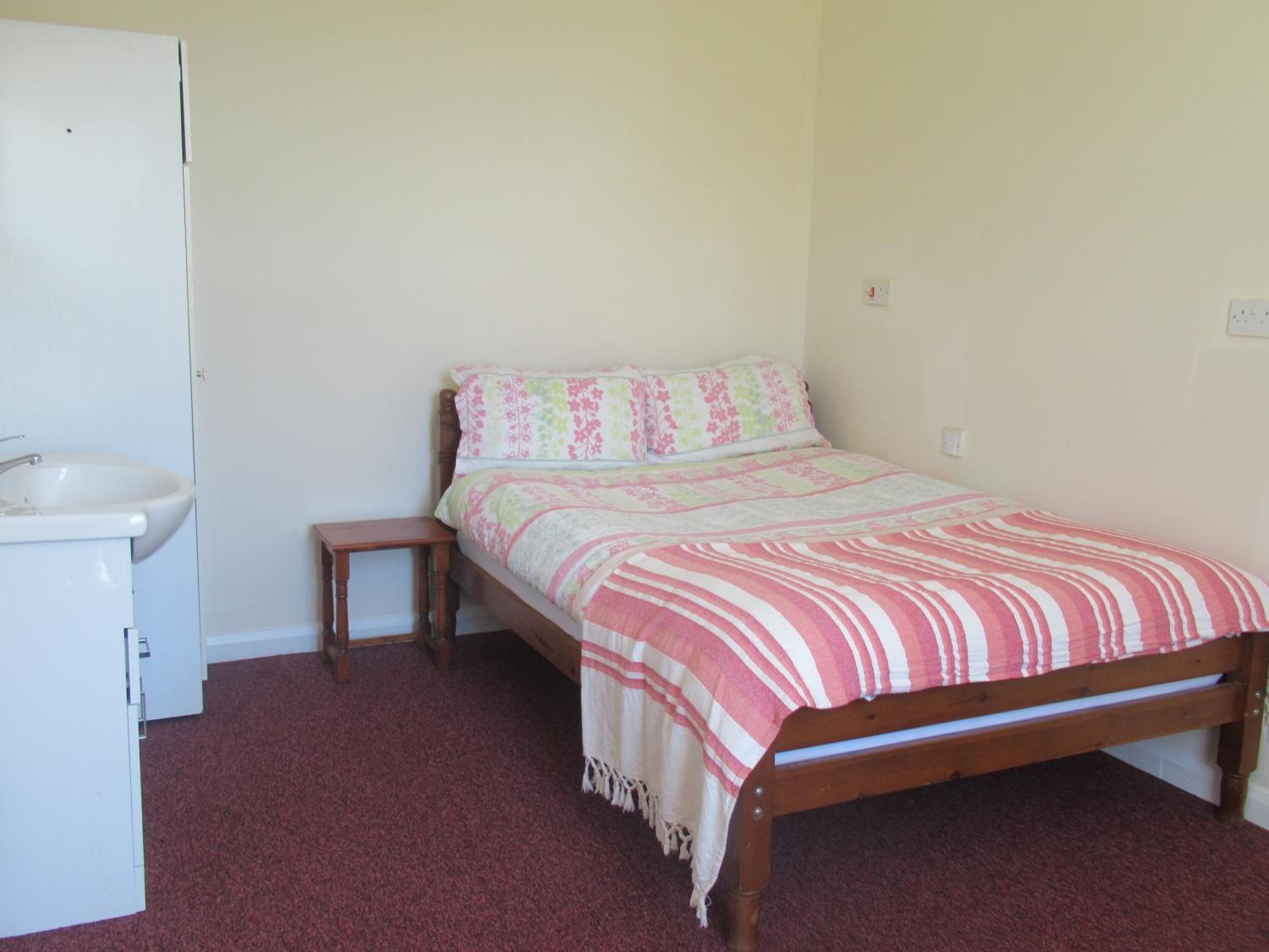A bedroom with a wooden bed, a bedside table, a white sink, and a red carpeted floor.