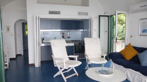 Un salon avec un sol bleu, des meubles blancs, un coin cuisine et une vue sur la verdure à l’extérieur.