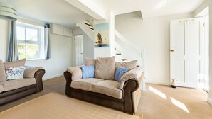 Un salon spacieux avec un canapé marron et un pouf assorti, un escalier blanc et une porte dotée d’un trou de serrure.
