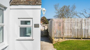 Une petite maison blanche avec un toit en tuiles, une clôture en bois et un ciel d’un bleu éclatant.