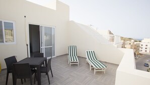 A balcony with a dining table and chairs, two striped lounge chairs, and a view of the cityscape. - Dolphin Court Gozo (Zebbug)