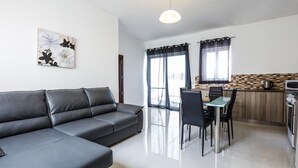 A modern living room with a grey sofa, a dining table with chairs, a kitchen area with a microwave, and a large window with curtains. - Dolphin Court Gozo (Zebbug)