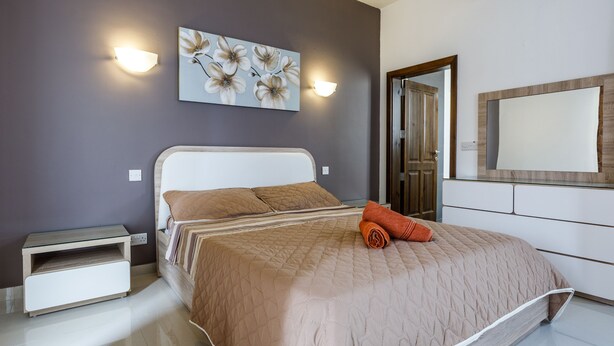 A bedroom with a bed, a nightstand, a wall-mounted light, a painting, and a mirror. - Dolphin Court Gozo (Zebbug)