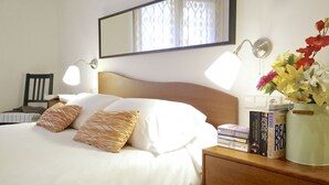 Un lit bien fait, avec du linge de lit blanc et des coussins à motifs, une table de chevet avec des livres et un vase de fleurs, et un miroir fixé au mur.