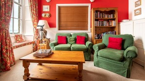 A cozy living room with a green sofa set, a wooden coffee table, a bookshelf, and a window with curtains. - Tittle Tattle - One Bedroom Cottage, Sleeps 2 (Penzance)