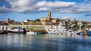A harbor with boats docked, a prominent church with a tower, and residential buildings in the background. - Tittle Tattle - One Bedroom Cottage, Sleeps 2 (Penzance)