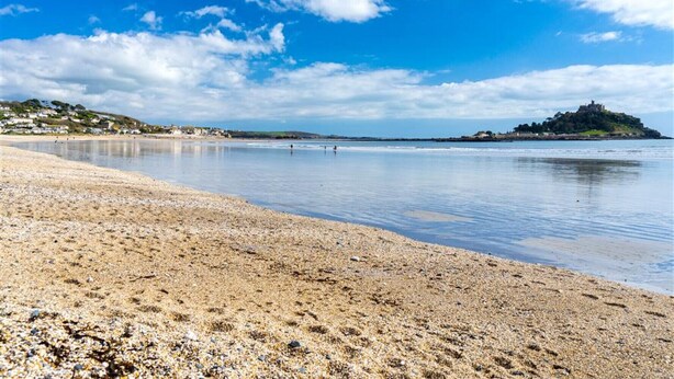 A beach with pebbles, calm water, and a distant island with buildings. - Tittle Tattle - One Bedroom Cottage, Sleeps 2 (Penzance)