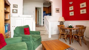 A cozy living room with a green sofa, a wooden coffee table, and a dining area with wooden chairs and a table. - Tittle Tattle - One Bedroom Cottage, Sleeps 2 (Penzance)