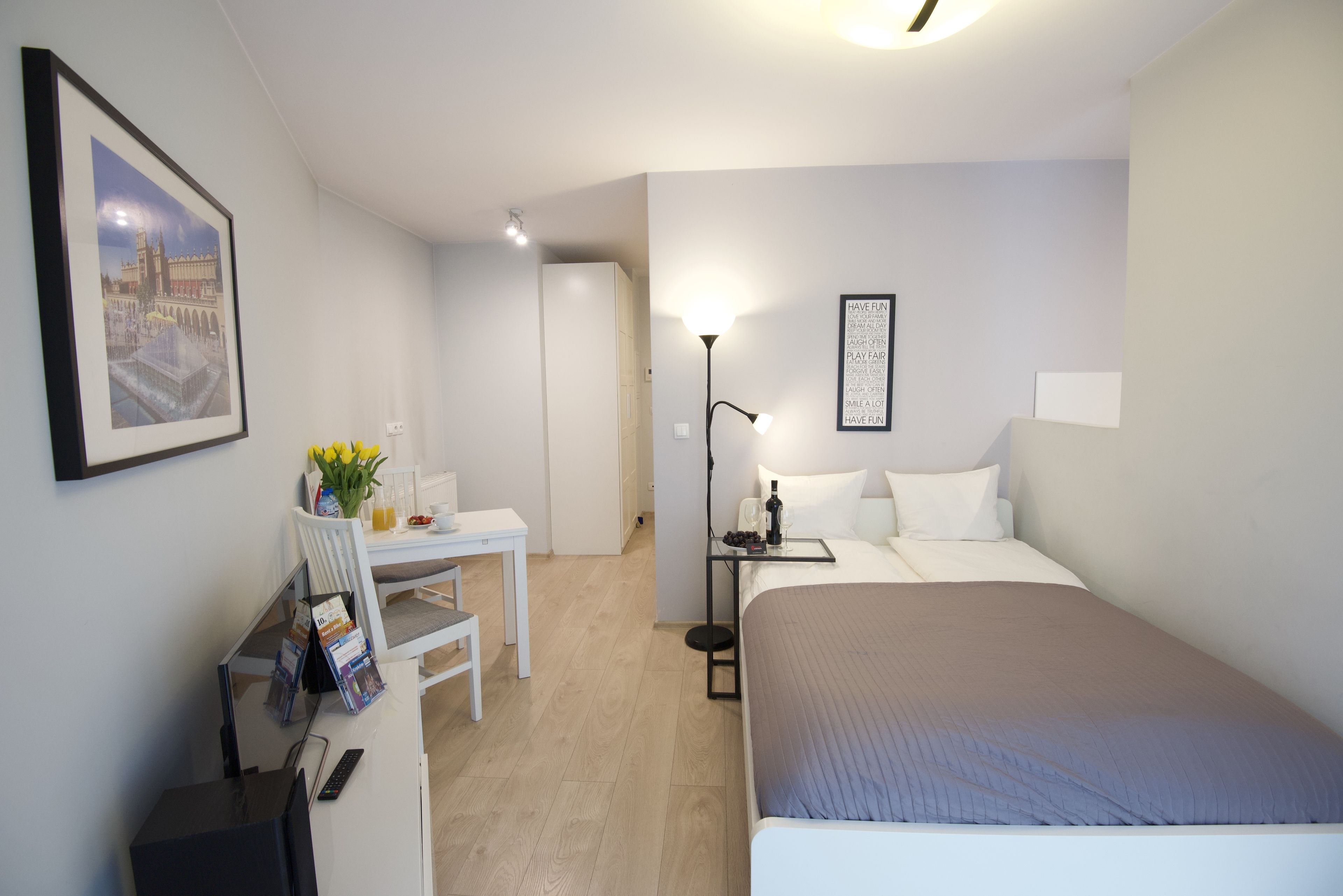 Habitación de hotel con cama, escritorio con computadora y silla, mesita de noche con lámpara, y un cuadro enmarcado en la pared.