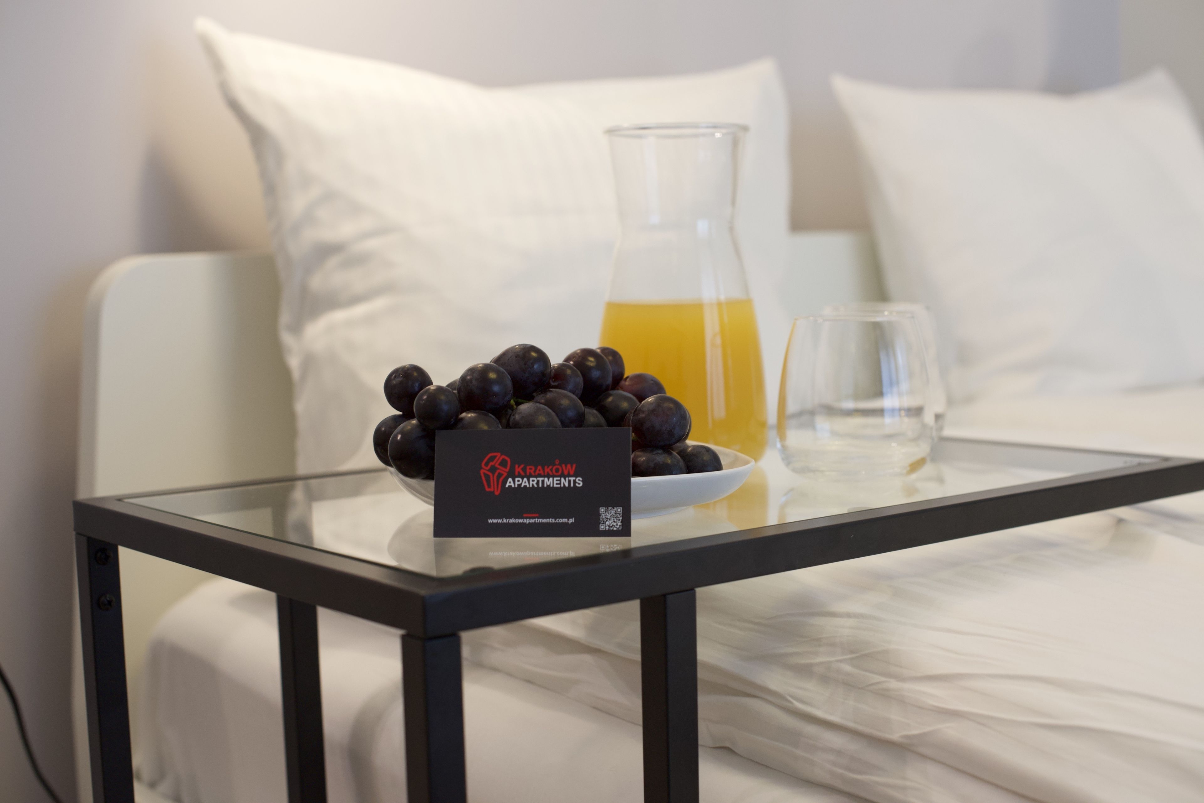 A glass table with a bowl of dark fruits, a glass pitcher of yellow liquid, and a glass on a bed with white pillows.