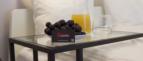 Una mesa de cristal con un bol de frutas oscuras, una jarra de cristal con líquido amarillo y un vaso sobre una cama con almohadillas blancas.