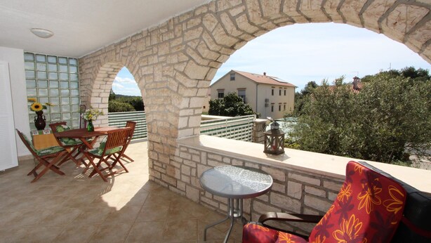 Un balcón con arcos de piedra, una mesa de comedor con sillas, una mesita redonda y vistas a casas y árboles.