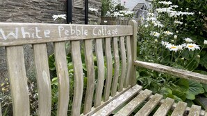 A wooden bench with "White Pebble Cottage" written on it, surrounded by greenery and white flowers.
