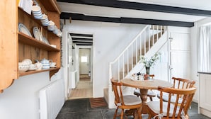 A dining area with a round wooden table and four chairs, a wooden shelf with dishes, and a staircase leading to another room.