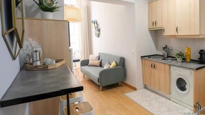 Une cuisine compacte avec un lave-linge, un coin repas avec une table et des chaises, et un canapé gris dans un espace de vie lumineux.