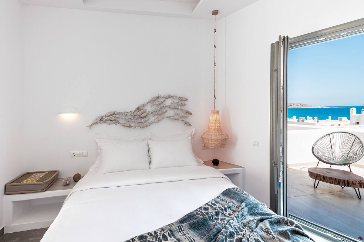 Ein Schlafzimmer mit Bett, Nachttisch, Lampe und Blick auf das Meer durch eine Glastür.