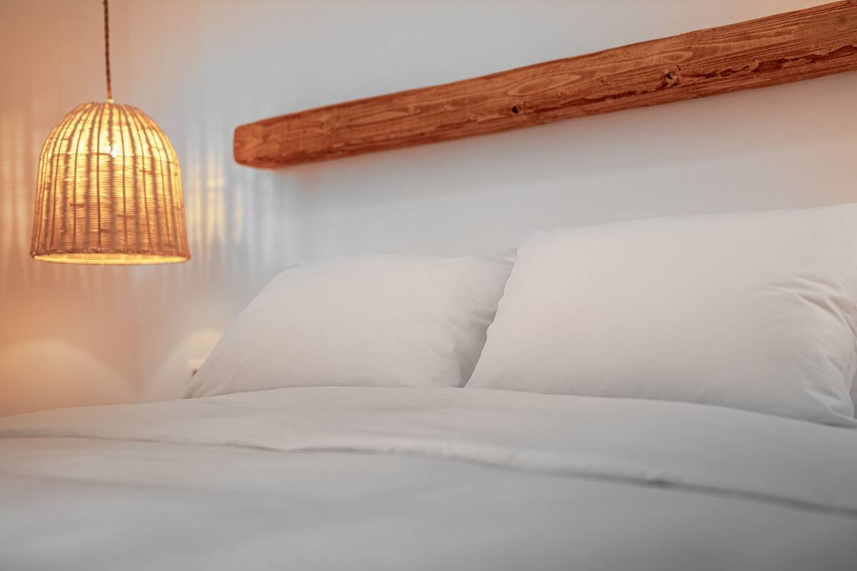 Ein Bett mit weißer Bettwäsche und einer gewebten Leuchte darüber.