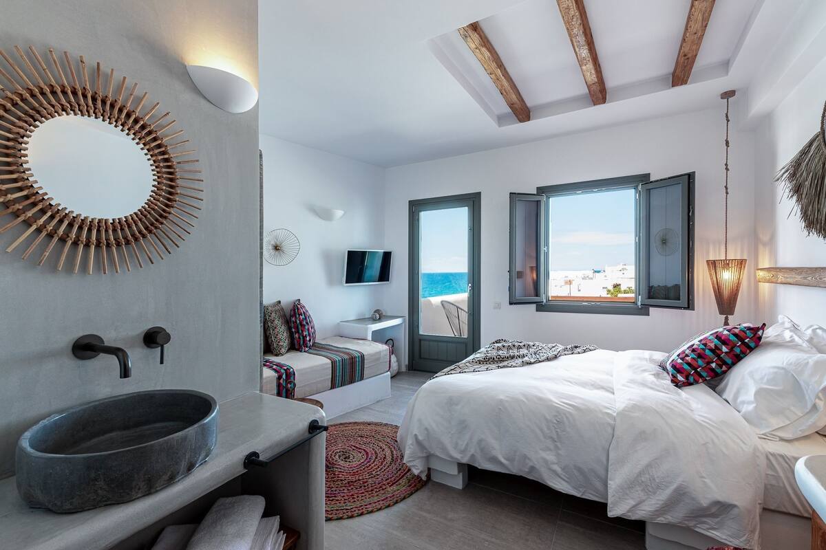 Ein modernes Schlafzimmer mit einem großen Bett, einem integrierten Waschbecken, einem Fernseher und Meerblick durch eine offene Tür.