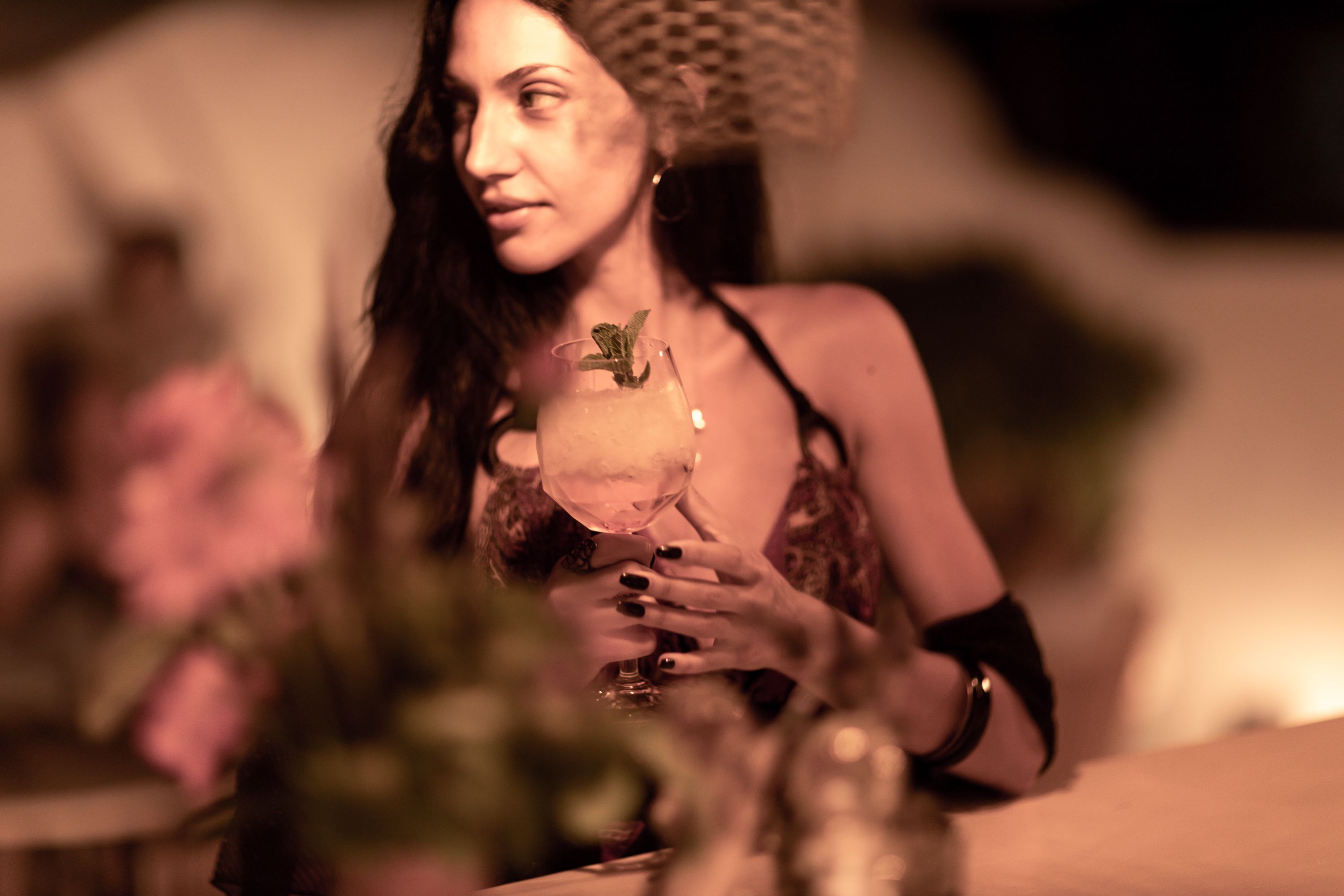 A person holding a glass with a mint garnish, seated at a table with a vase of flowers.