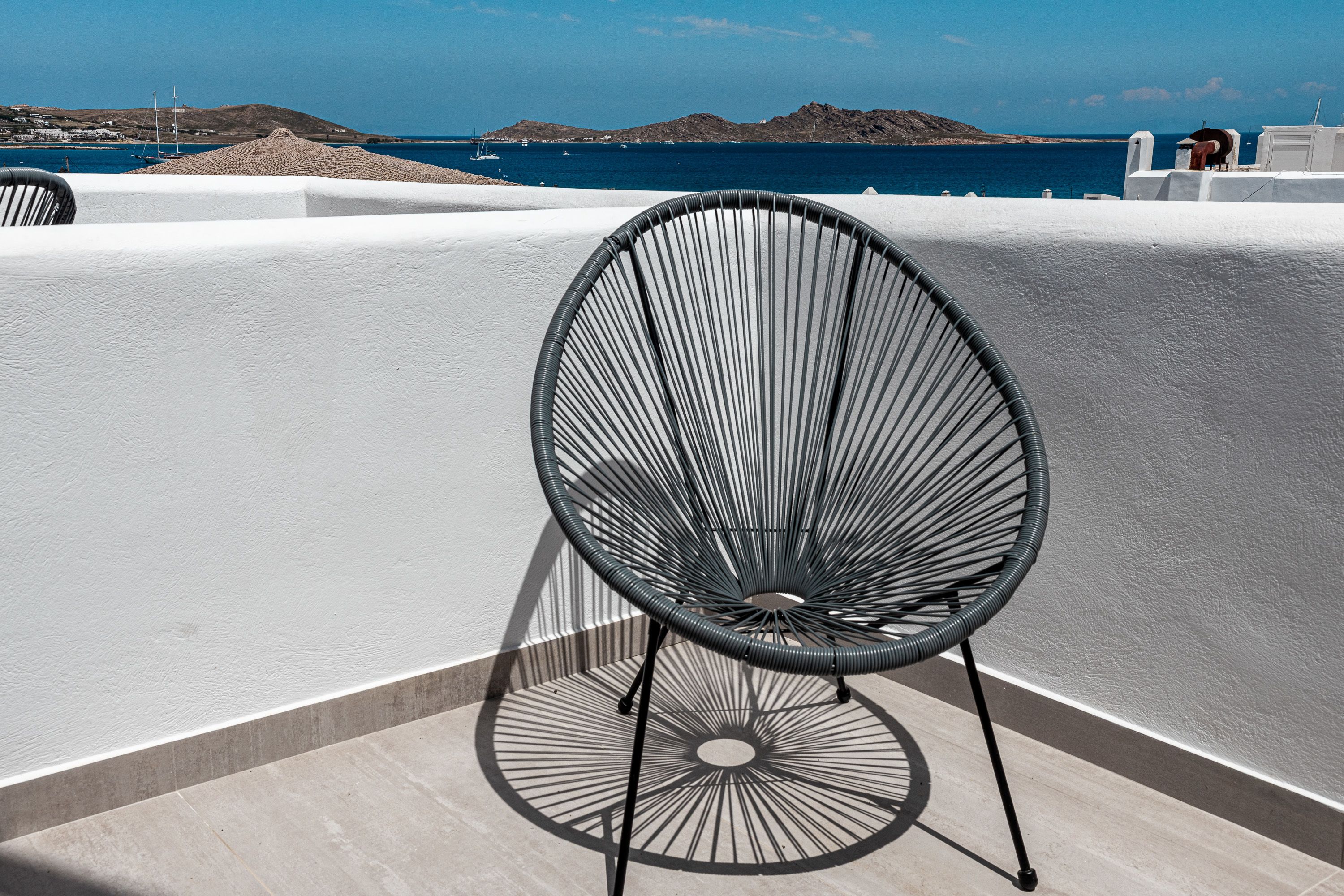 A black wireframe chair on a balcony with a white wall and a view of the sea and hills.
