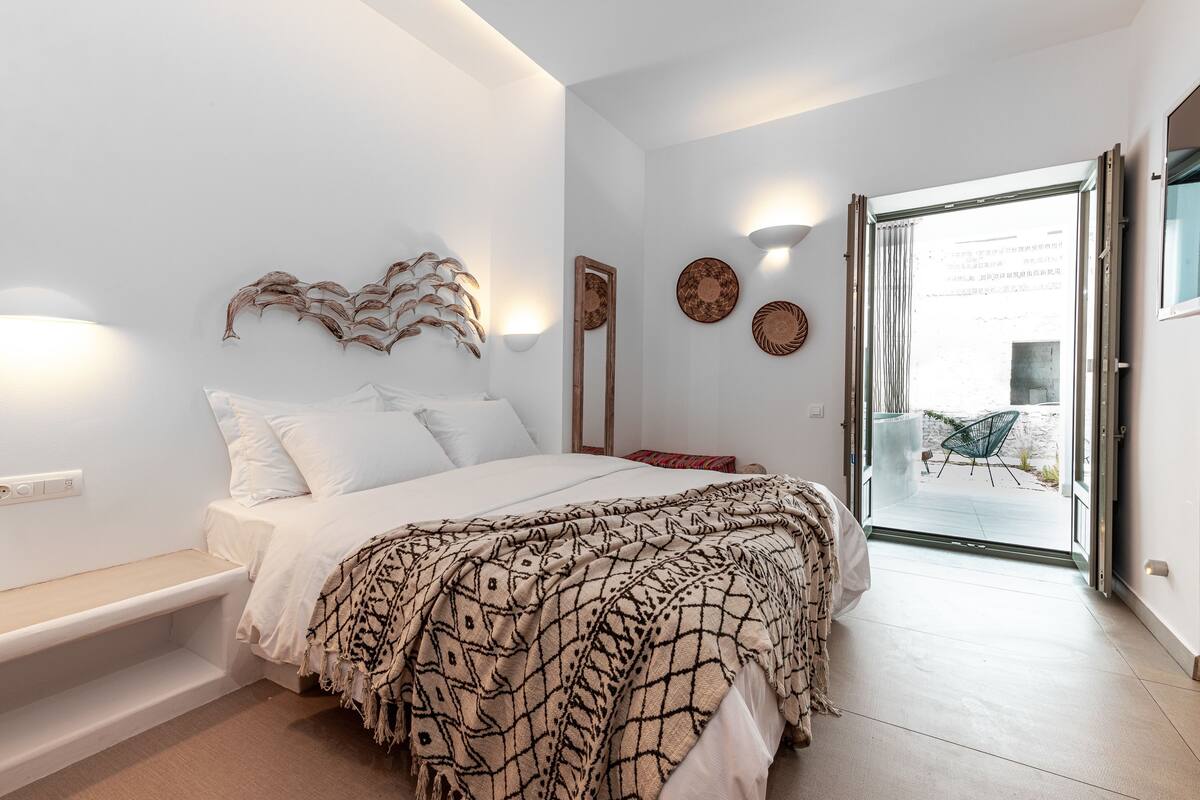 Ein Schlafzimmer mit einem Bett, einem Nachttisch und einer Bank. Ein Fenster mit Vorhängen bietet einen Blick nach draußen.