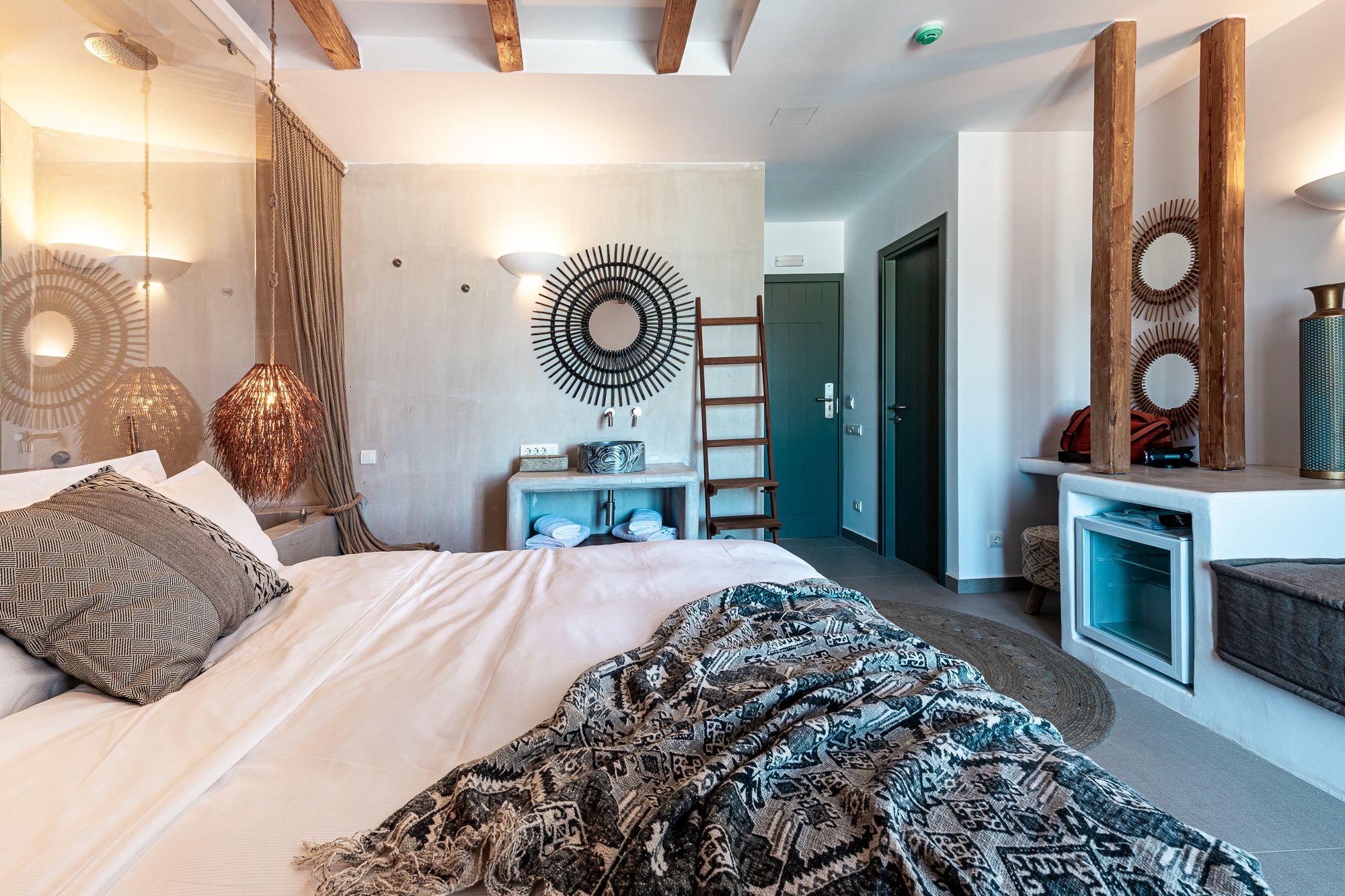 A modern bedroom with a large bed, a wall-mounted circular mirror, a ladder, and a small table with a lamp.