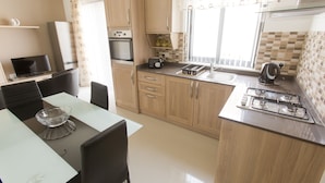 A compact kitchen with wooden cabinets, a gas stove, a microwave, and a small dining area with a glass table and chairs. - Waterside Apartments (Zebbug)