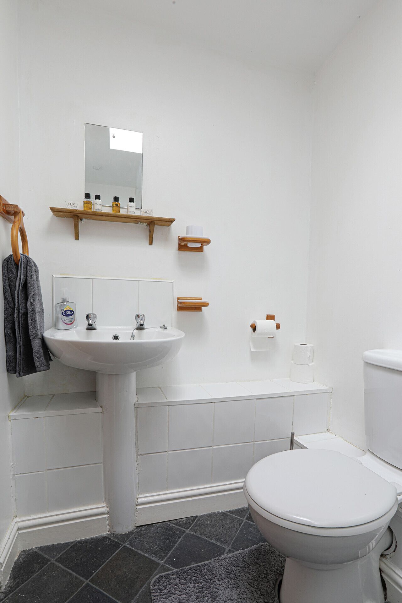 Un bagno con lavandino bianco, specchio e servizi igienici. Si nota un portasciugamani, uno scaffale con articoli da toilette e un porta carta igienica.