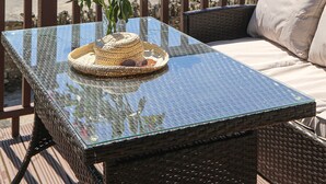 Une table à plateau en verre, des chaises en osier et un coussin, un panier en osier et un vase avec des fleurs rouges, le tout sur un balcon donnant sur un paysage pittoresque.