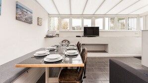 Et spiseområde med et langt bord dækket til seks personer, et fjernsyn ophængt på væggen og et vindue med udsigt til det fri.