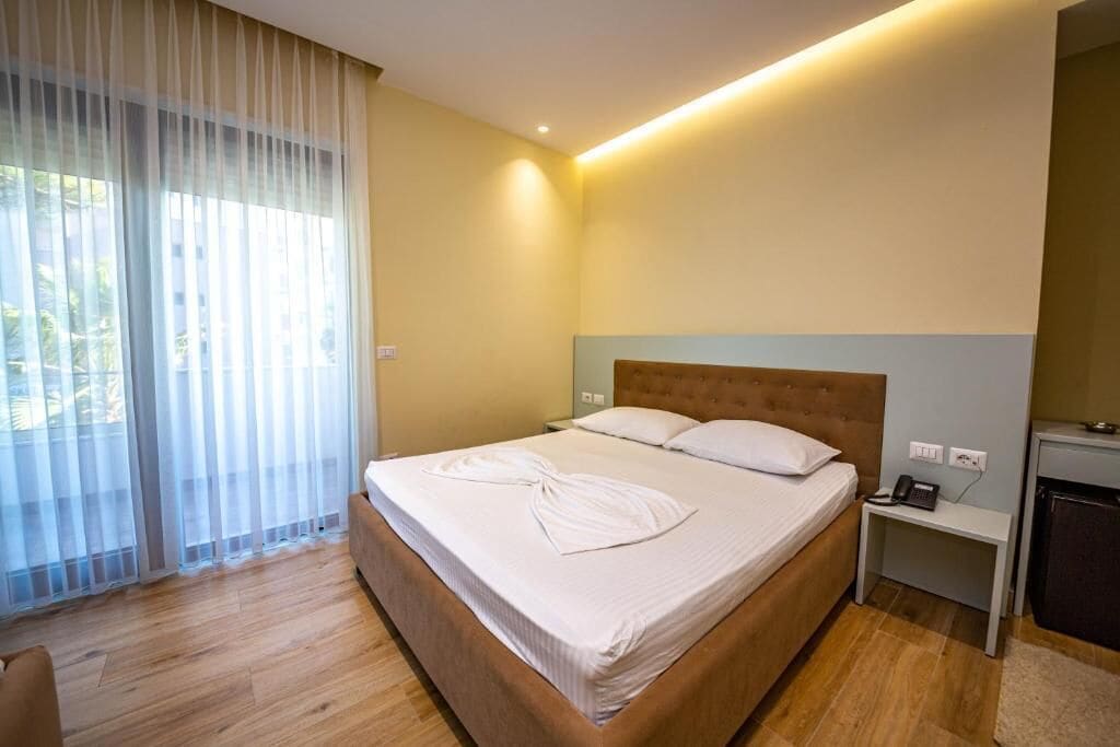A neatly made bed with a wooden headboard, a bedside table with a phone, and a window with sheer curtains.