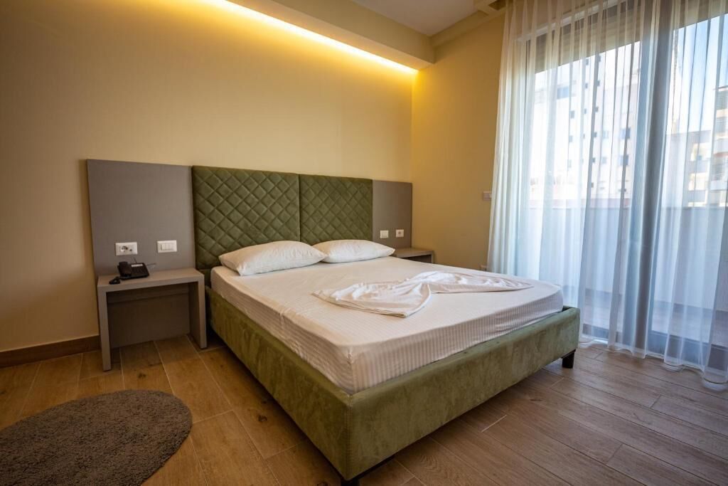 A hotel room with a green upholstered bed, two bedside tables, a bedside lamp, and a view through sheer curtains.