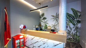 Un área de recepción con mostrador de mármol, máquina expendedora roja y papel tapiz tropical.