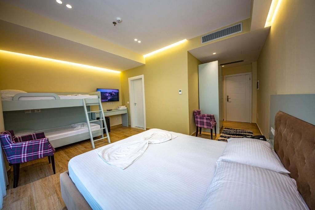 A hotel room with a bunk bed, a single bed, a television, a chair, and a door leading to another room.
