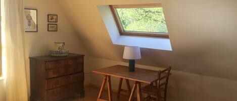 Een kleine, knusse kamer met een schuine kap, een houten bureau met een lamp, een stoel en een raam met uitzicht op groen.