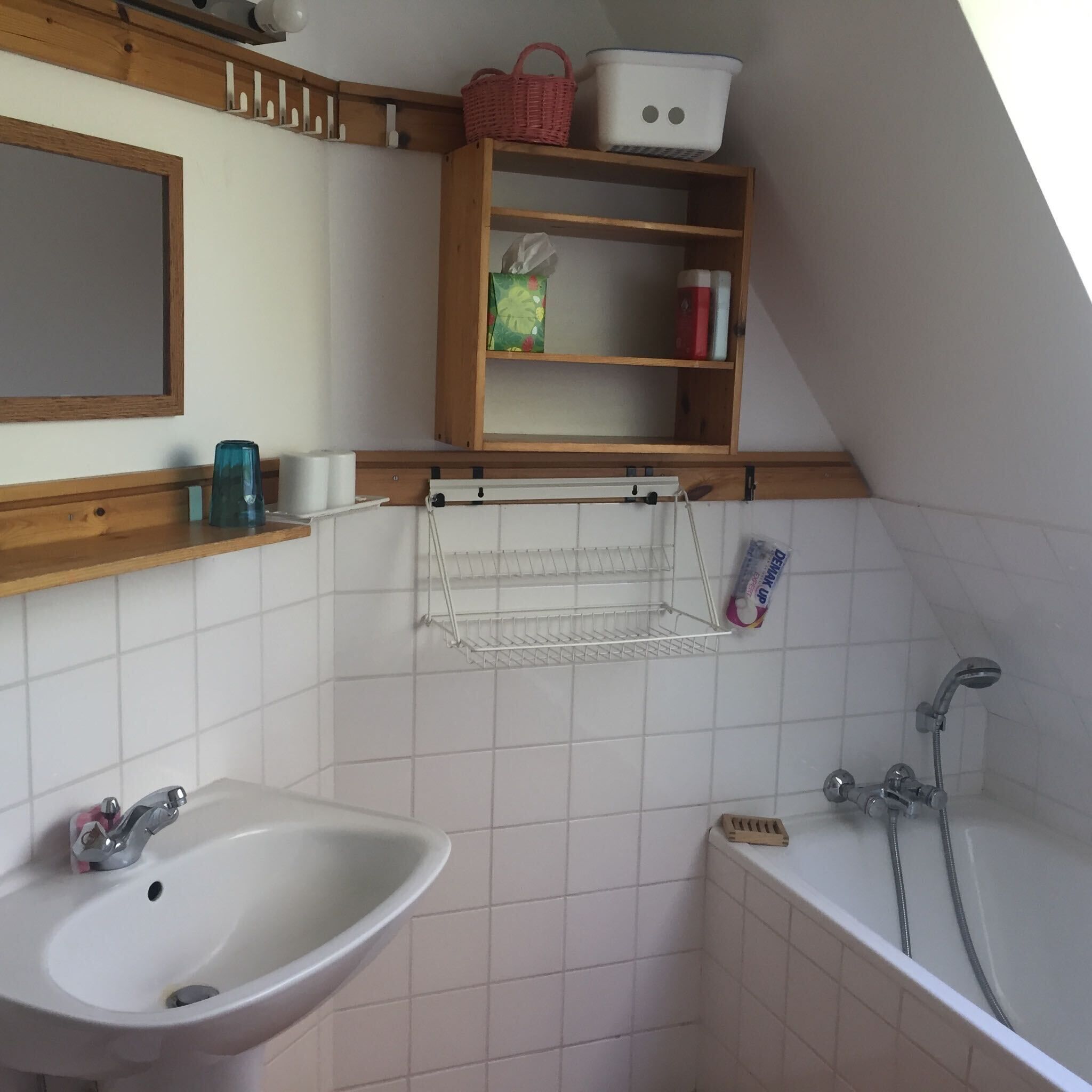 Uma casa de banho com uma pia branca, um espelho, uma prateleira com artigos de higiene pessoal, um chuveiro e um porta-toalhas.