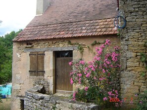 Un edificio rústico de piedra con tejado de baldosas, una puerta de madera y plantas florecientes de color rosa al frente.