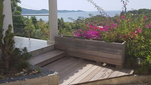 Een houten terras met een bankje, omringd door planten en uitzicht op zee.