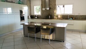 Een moderne keuken met witte kasten, een groot eiland met twee stoelen, roestvrijstalen apparatuur en een raam dat natuurlijk licht binnenlaat.
