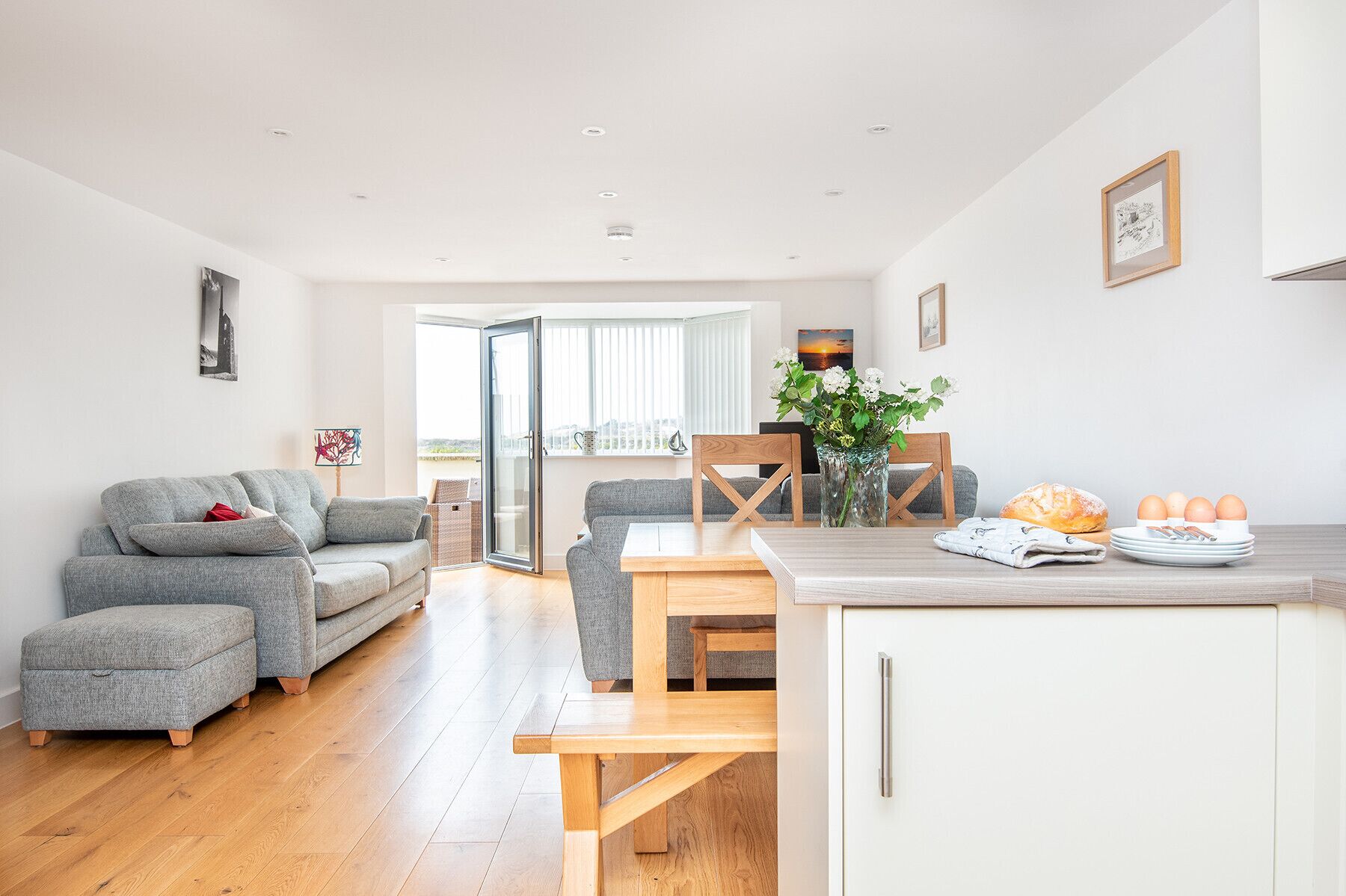 Un espacio moderno para vivir con un sofá gris, una mesa de comedor de madera y una zona de cocina con pan, huevos y un florero.