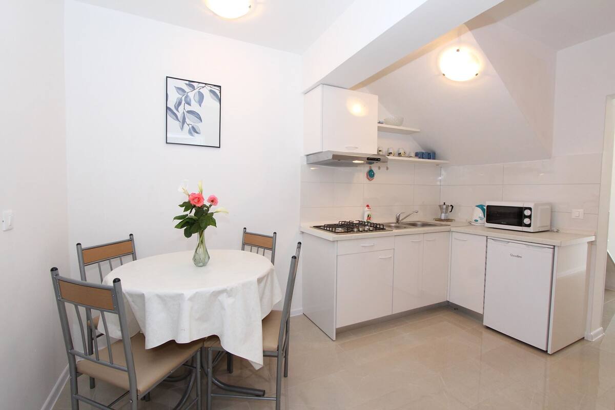 Une cuisine compacte avec des meubles blancs, un four à micro-ondes et une cuisinière à gaz. On y trouve une table à manger avec des chaises et un vase rempli de fleurs.