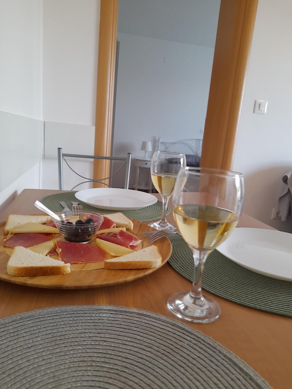 A dining table with a plate of cheese and ham, two glasses of white wine, and a bowl of olives.