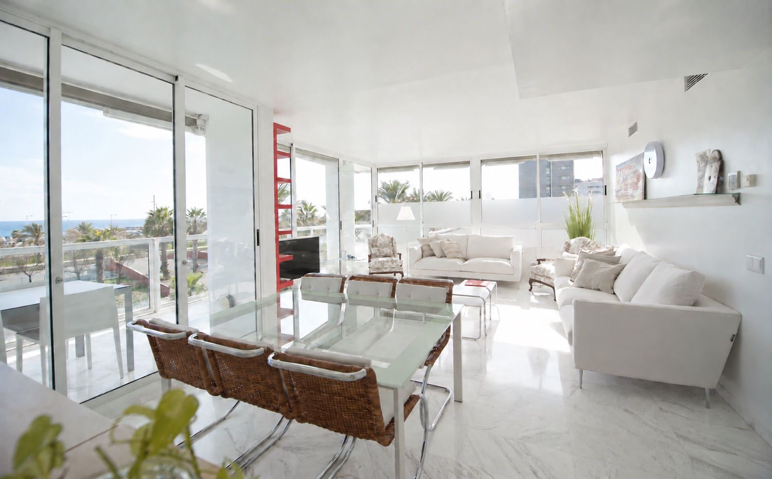 A modern living room with a glass dining table, white sofas, and a balcony with a view.