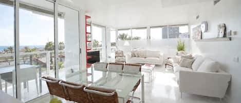 A modern living room with a glass dining table, white sofas, and a balcony with a view.