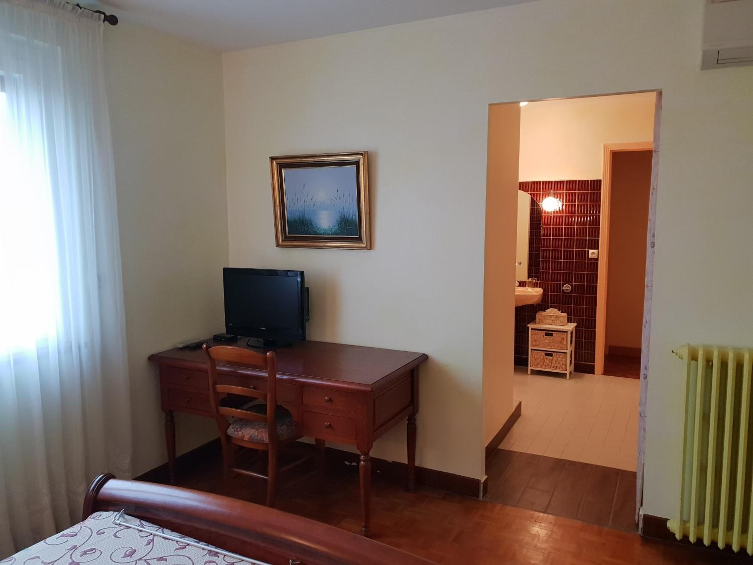 Una habitación con un escritorio de madera, una silla, un cuadro en la pared y vistas a un baño a través de un vano abierto.