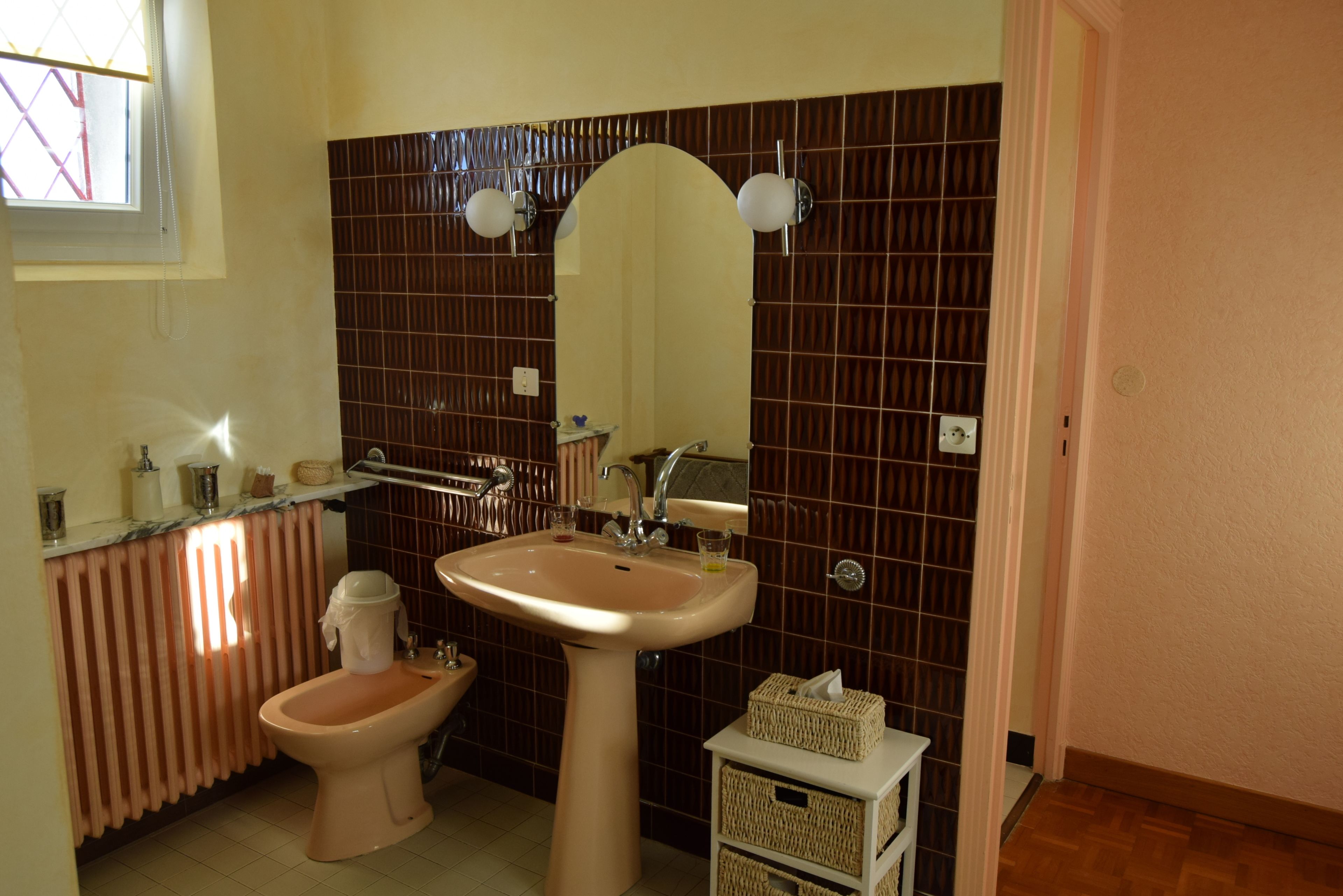Une salle de bain équipée d’un lavabo, d’un bidet et d’un miroir. On y trouve une fenêtre avec des stores, un radiateur et un petit panier posé sur le comptoir.