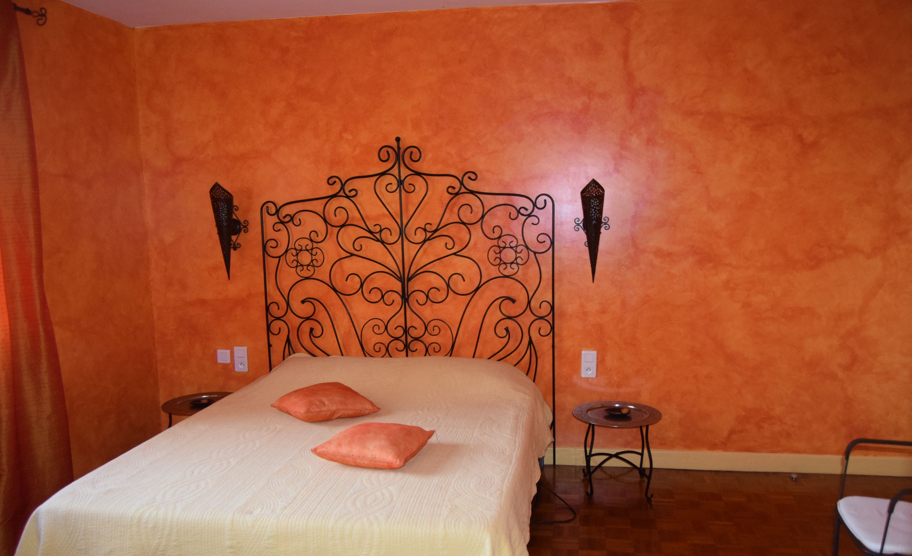 A bedroom with an orange wall, a decorative metal headboard, two pillows, a small round table, and a chair.