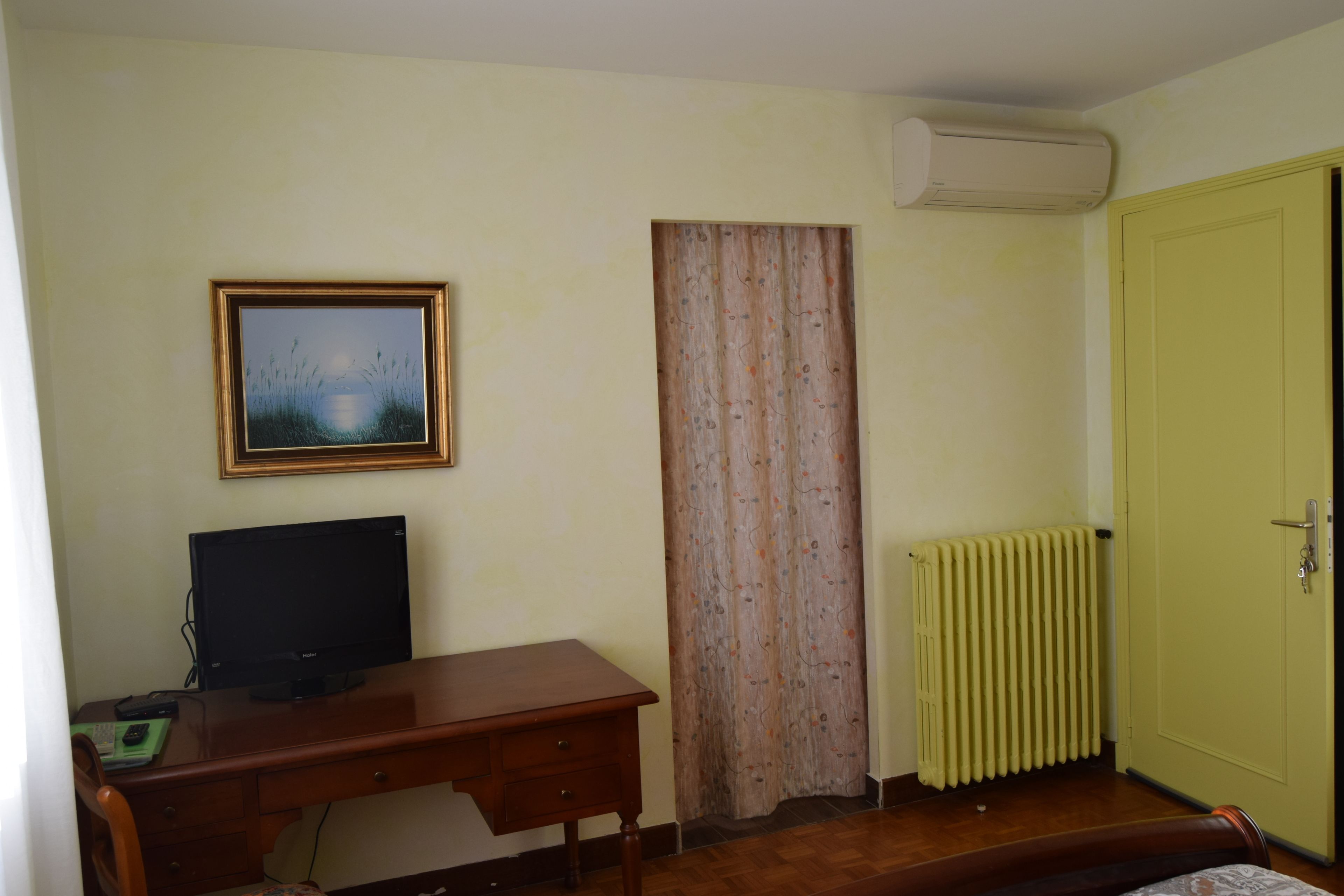 Une pièce avec un bureau en bois, un écran noir, un tableau encadré, une fenêtre avec un rideau, un radiateur et une porte verte.
