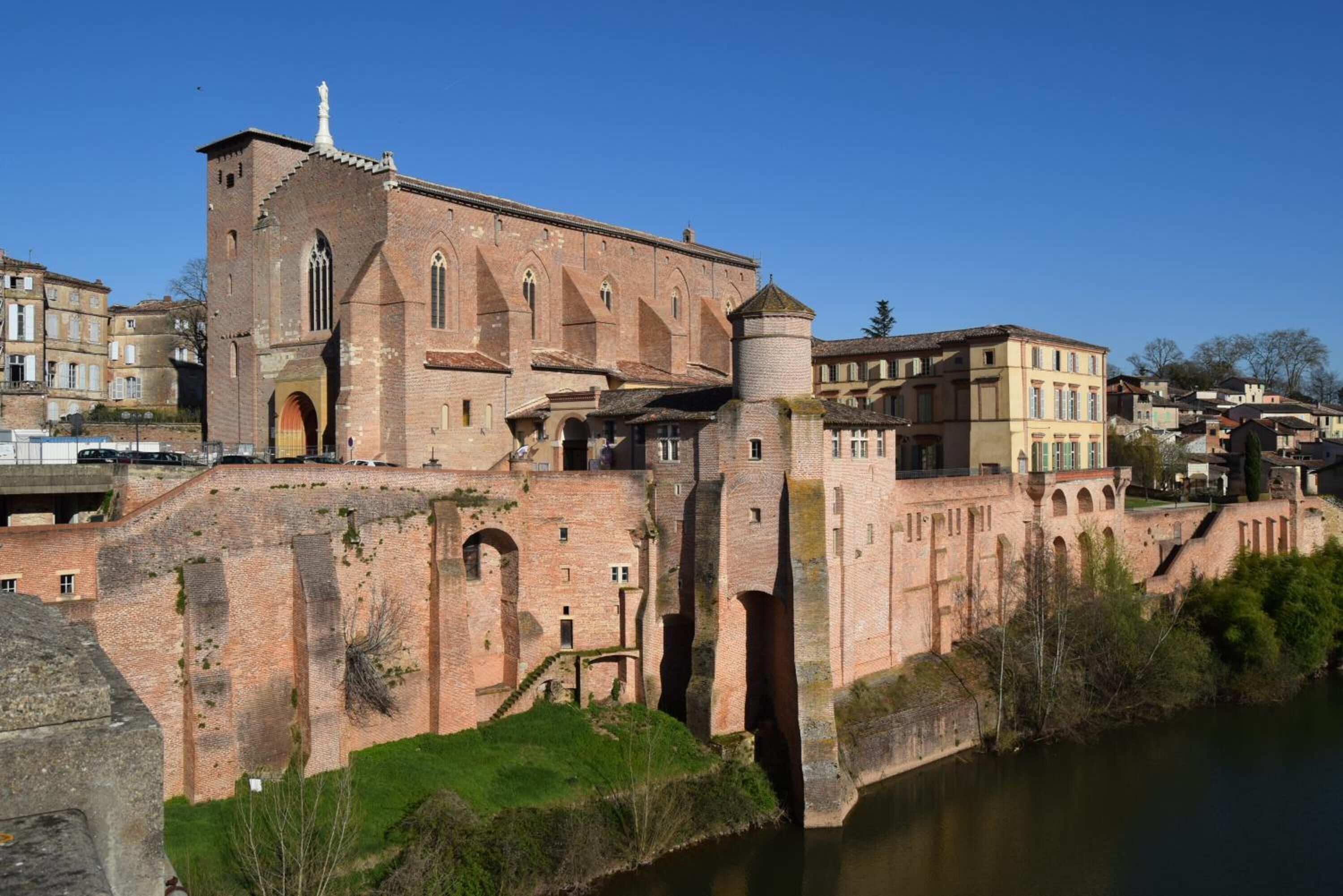 Un edificio histórico con una torre destacada, ventanas de arco y una muralla fortificada que da vista a un río.