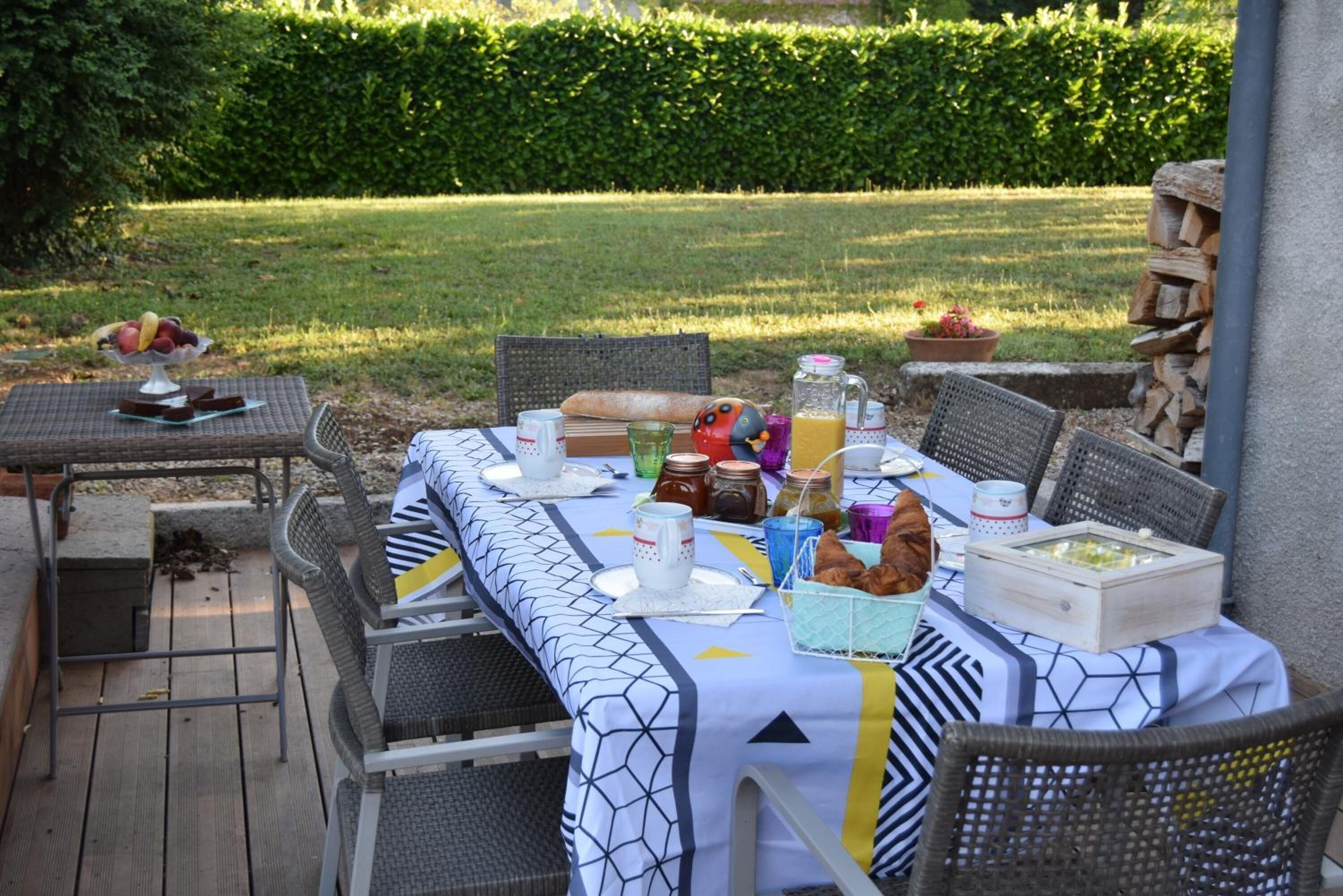 An outdoor dining area with a table set for a meal, surrounded by wicker chairs, and a view of a well-maintained lawn and hedge in the background.